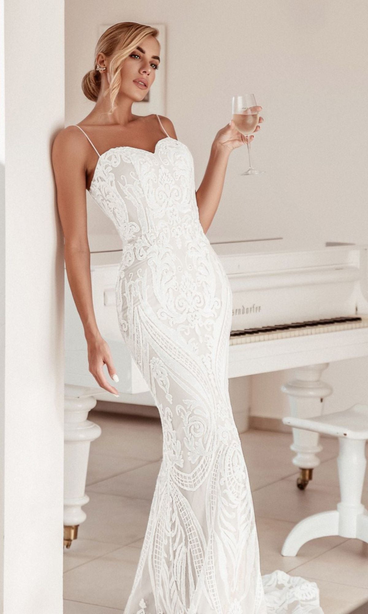 A woman in the White Lace Bodycon Bridal Gown: Tina Holly BA999 stands next to a white piano, holding a glass of wine and gazing away from the camera.